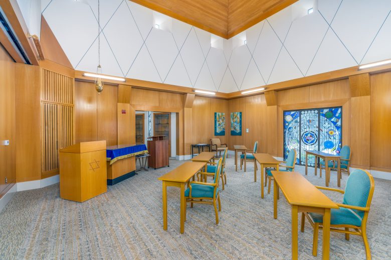 A small chapel with wooden walls, several tables and chairs, a podium, a piano, and stained glass windows with blue designs. The room is well-lit with both natural and artificial light.